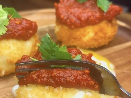 Round golden fritters topped with red sauce, white cheese, and fresh cilantro on wooden board